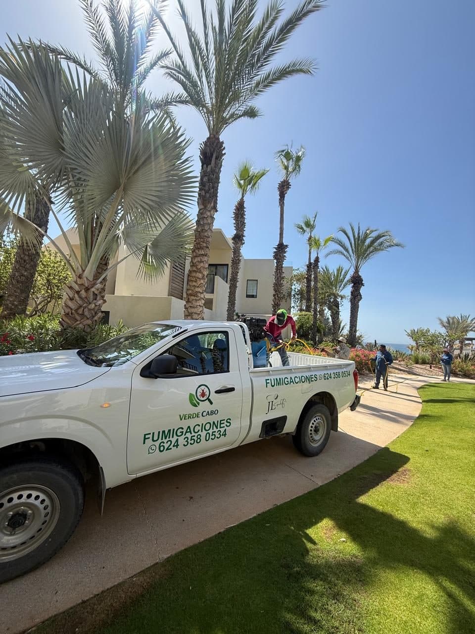 Camioneta de Verde Cabo con equipo de fumigación, entre palmeras y pasto cuidado, con el mar al fondo